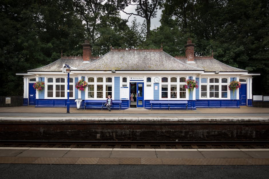 A scenic view of a charming train station building painted in white and blue, with multiple large windows and hanging flower baskets filled with colorful blooms. The station features a central waiting