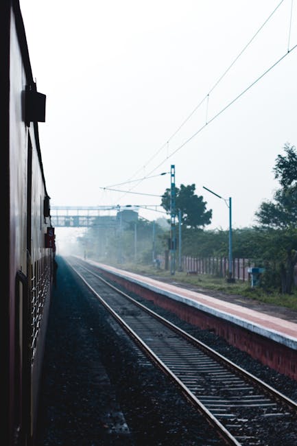 This image shows a view from a train carriage window, capturing the railway tracks extending into the distance beneath overhead electric wires. The scene is set on a misty day with soft, diffused ligh