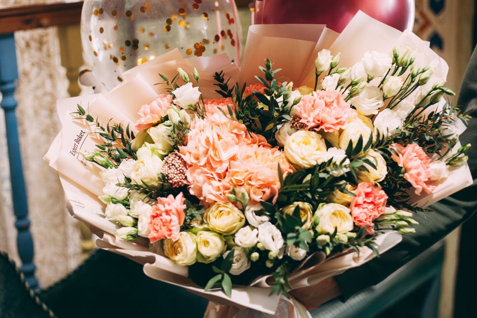 A vibrant floral bouquet featuring a mix of peach, white, and cream-colored roses, carnations, and lisianthus, accented with dark green foliage. The flowers are arranged in a rounded, slightly cascadi